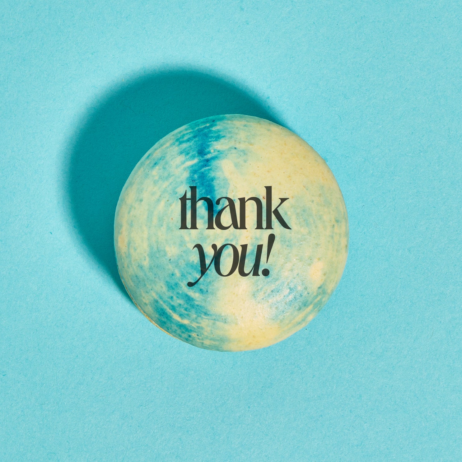 Overhead shot of a yellow and blue tie-dye macaron with the words "thank you!" printed in black ink on top. The macaron is placed on a bright aqua backdrop.