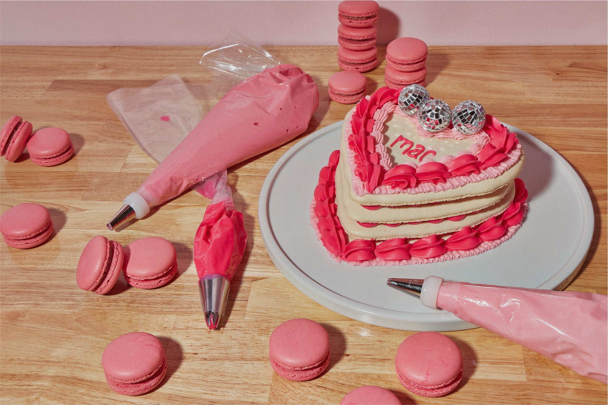 One of Maddie About Cake's signature heart-shaped macaron cakes, complete with pink icing and mini disco balls, surrounded by piping bags of icing and strawberry macarons.