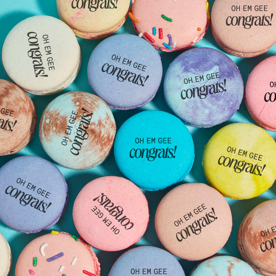 Overhead shot of various macarons colors and flavors printed with the words "OH EM GEE congrats!" in black text.