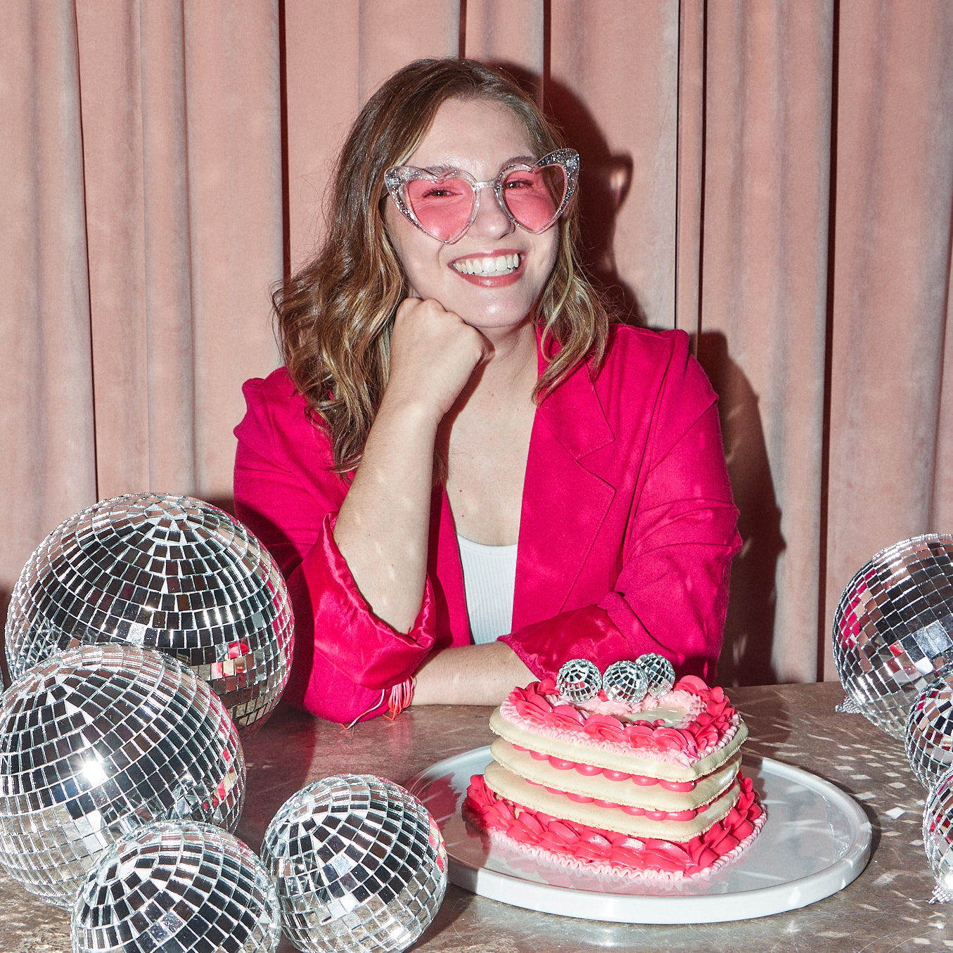 Maddie, founder and head baker at Maddie About Cake, is smiling and posting with one of her signature heart shaped macaron layer cakes, surrounded by different sized disco balls. Maddie is wearing a hot pink blazer and sparkly, pink heart shaped sunglasses.