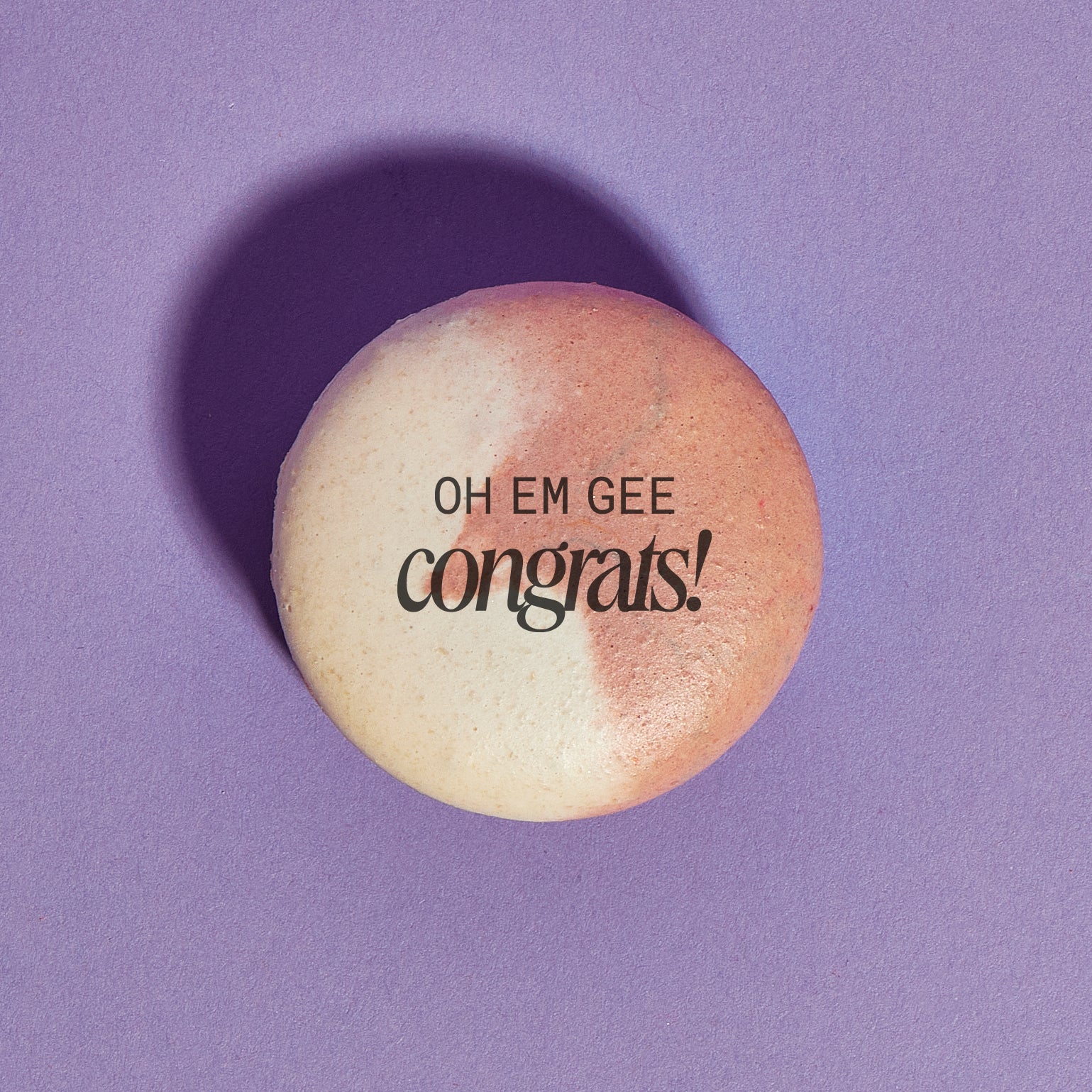 Overhead shot of a brown and white swirled macaron with the words "OH EM GEE Congrats!" printed on the shell in black ink. The macaron is placed on a purple backdrop.