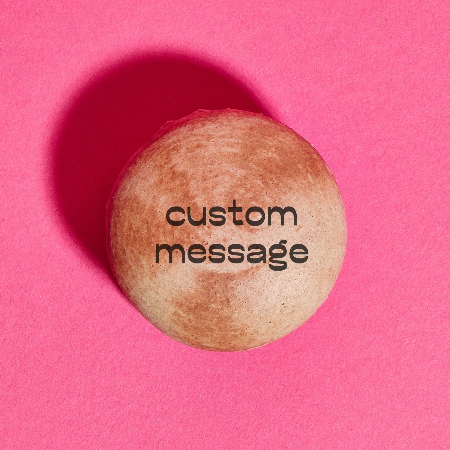 Overhead shot of a brown tie-dye macaron with the words "custom message" printed on the shell in black ink. The macaron is placed on a hot pink backdrop.