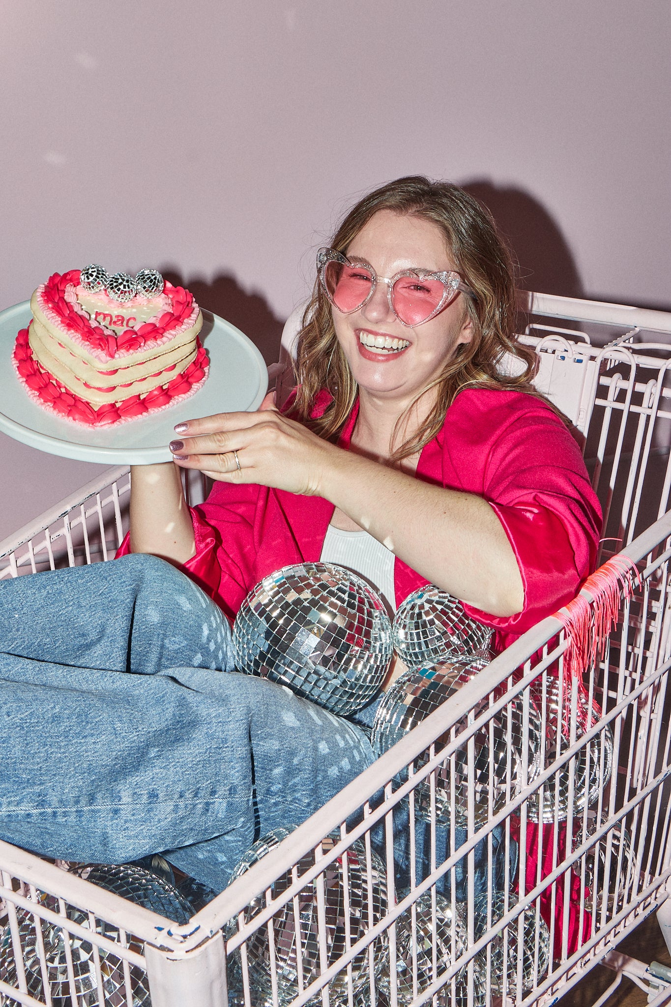 Maddie, owner and head baker of Maddie About Cake, is sitting in a light pink shopping cart surrounded by disco balls and holding a pink heart-shaped macaron cake topped with mini disco balls. She is smiling at the camera and wearing light pink heart-shaped glasses, a hot pink blazer with fringe, and blue jeans.