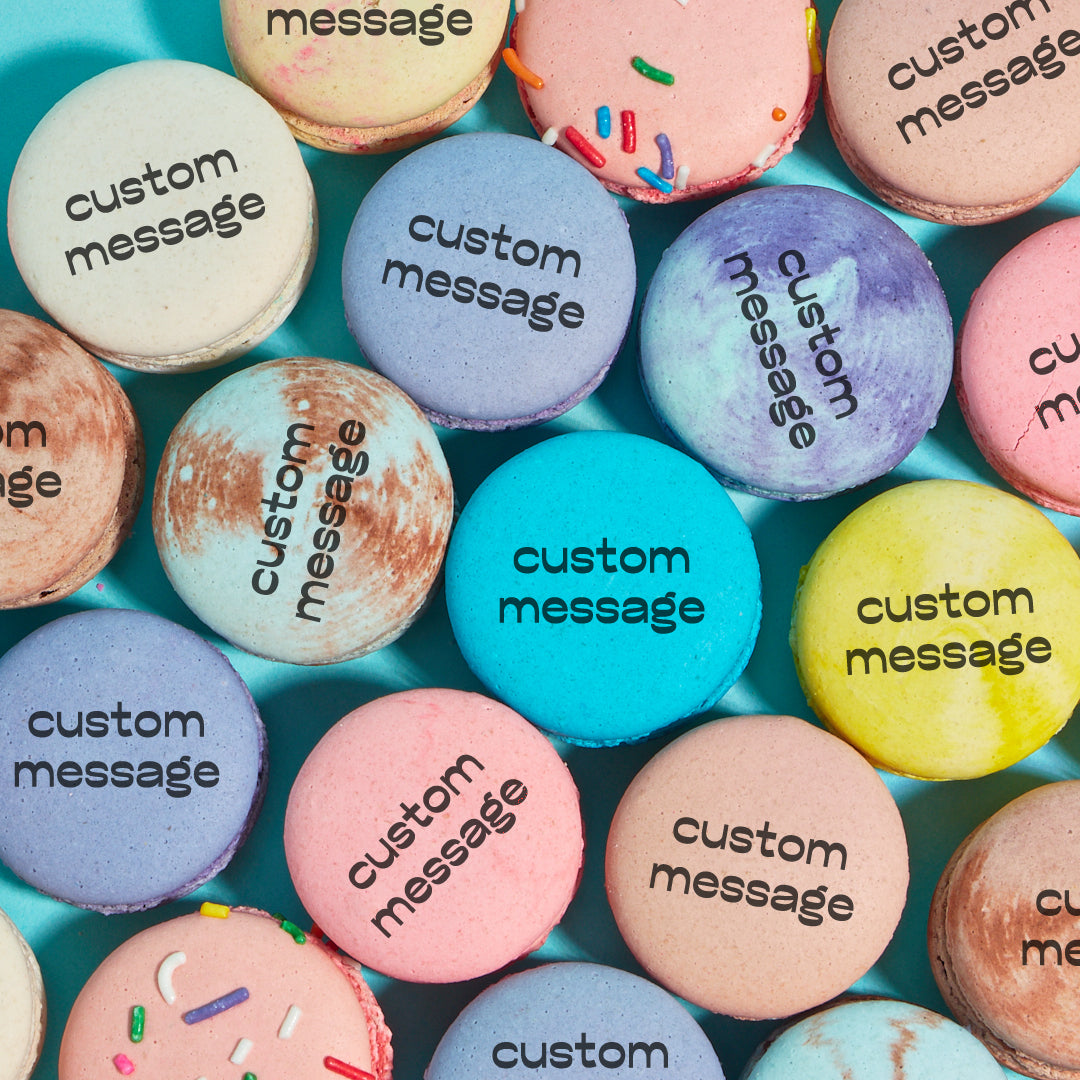Overhead shot of various macarons colors and flavors printed with the words "custom message" in black text, indicating that you can order macarons from Maddie About Cake with a custom message printed on them.