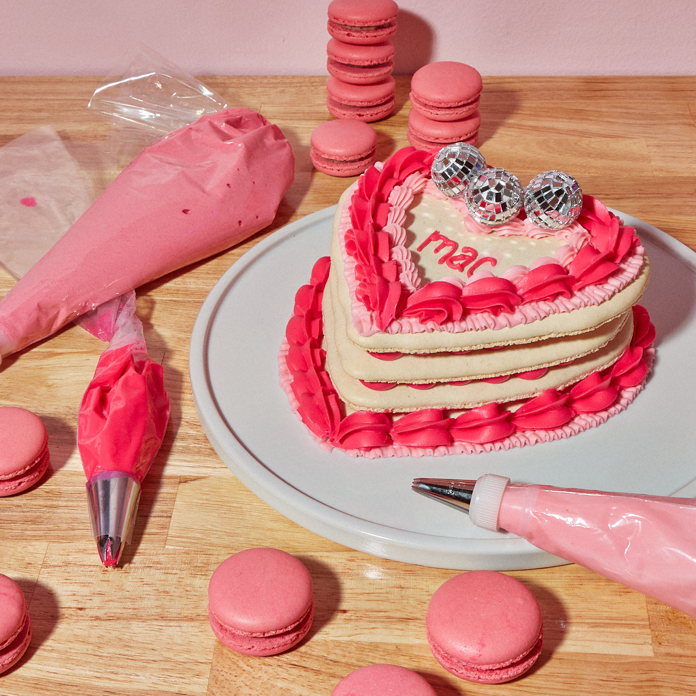 One of Maddie About Cake's signature heart shaped macaron layer cakes, complete with different shades of pink icing, custom text, and mini disco balls. The cake is surrounded by pink, strawberry macarons and three piping bags of pink icing.