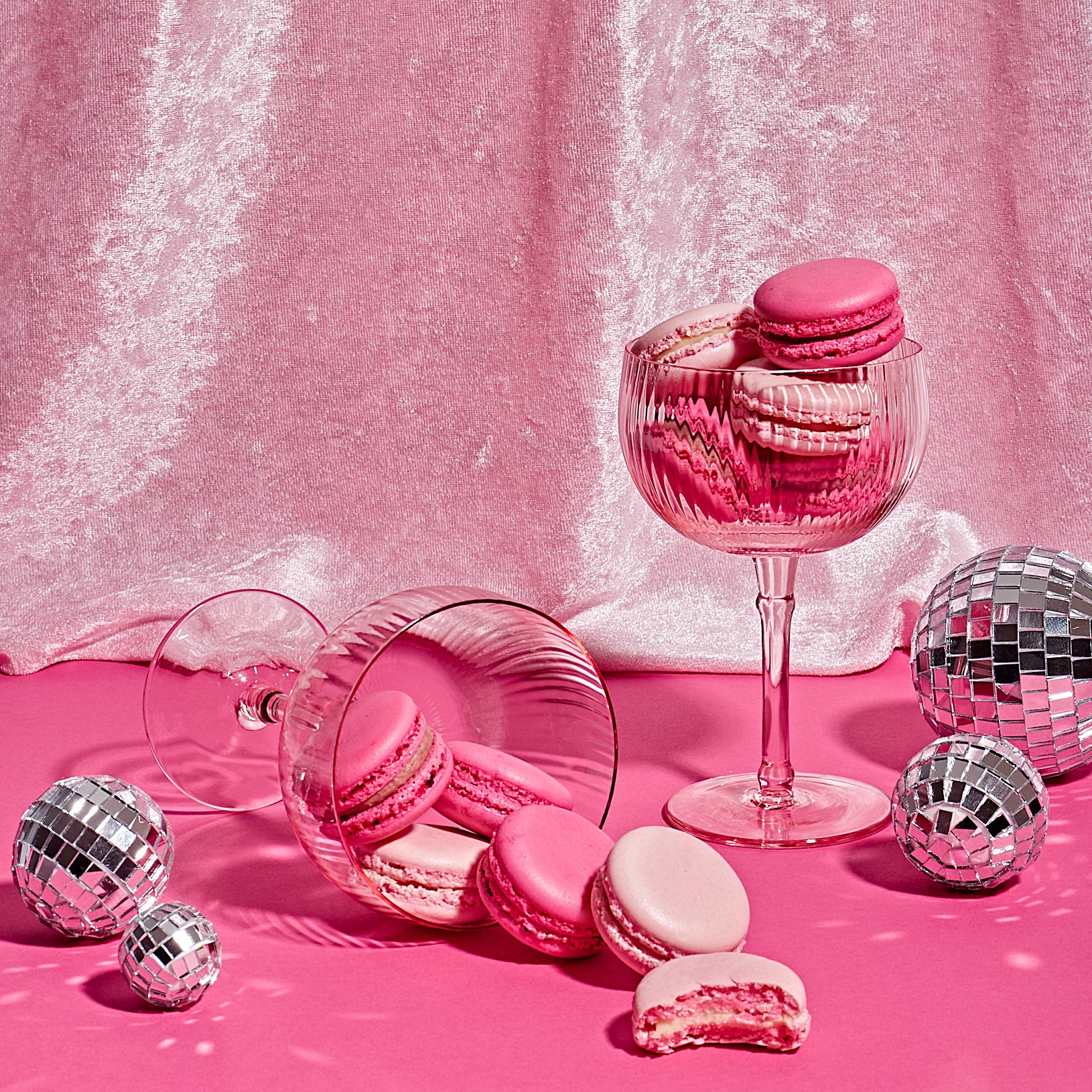 Two pink coupe glasses filled with various shades of pink, Valentine's themed macarons surrounded by small disco balls and placed on a pink backdrop. The coupe glass on the left has spilled, leaving a trail of macarons. The coupe glass on the right is upright and filled to the brim with macarons.
